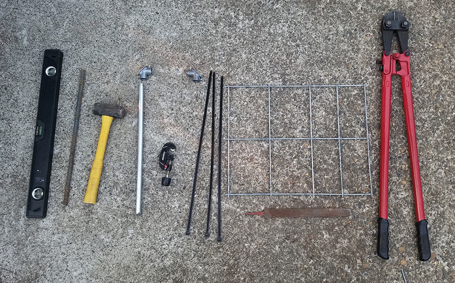 Tools and materials needed to make the trellis - left to right: level, rebar, big hammer, EMT with pull elbows above it, pipe cutter, hog wire with file underneath it, and huge bolt cutter
